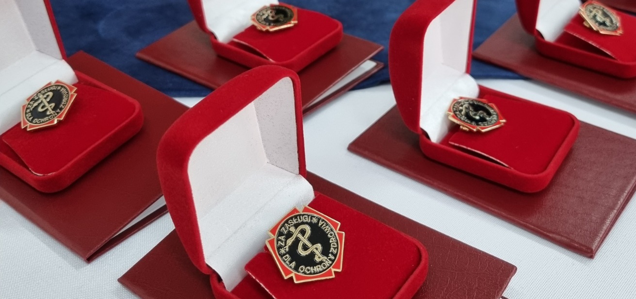 Na zdjęciu znajdują się odznaki "Za zasługi dla ochrony zdrowia" w eleganckich czerwonych pudełkach.