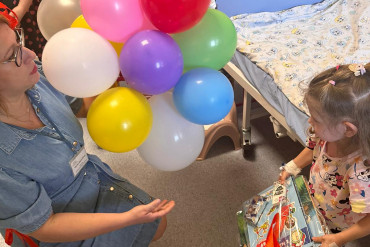 Kobieta z kolorowymi balonami kuca na przeciwko małej dziewczynki, która opiera się o łóżko szpitalne.