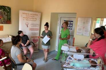 Pani dietetyk i położna stoją zwrócone w kierunku sedzących pacjentek. Po prawej stronie stoją Panie z noworodkami, które leżą wwózkach.