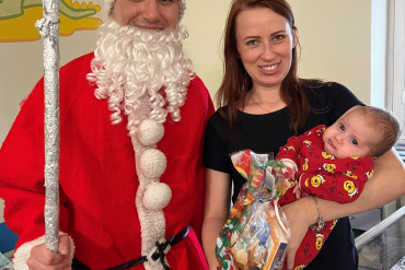 Na zdjęciu stoi mama trzymająca małe dziecko, obok nich stoi święty Mikołaj