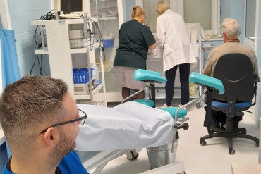 Na pierwszym planie siedzi lekarz przy biurku. W nieznacznej oddali stoją 2 pielęgniarki przygotowujące się do zabiegu. Obok nich na fotelu siedzi pacjent.