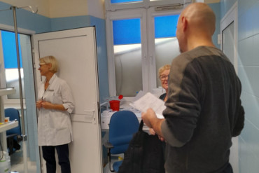 Pacjent stoi, trzymając w ręku dokumenty. Jedna z pielęgniarek uśmiecha się do niego, druga stoi w drzwiach i patrzy w przeciwną stronę.