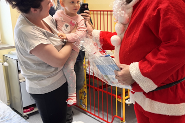 Mama trzyma na rękach dziecko, w ich kierunku pochyla się pracownik przebrany za św. Mikołaja