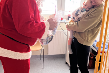 Mama trzyma na rękach dziecko, które się do niej przytula. Na przeciwko nich stoi św. Mikołaj trzymając w ręku prezent.