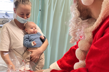 Mama trzyma na rękach dziecko, na przeciwko nich pracownik przebrany za św. Mikołaja wręcza na ręce mamy prezent dla dziecka.