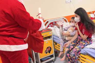 Mama siedzi na łóżku z dzieckiem, dziecko trzyma w rękach prezent, św. Mikołaj stoi na przeciwko nich