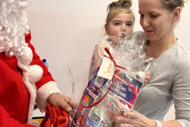 Mama trzyma na rękach dziecko, na przeciwko nich pracownik przebrany za św. Mikołaja wręcza na ręce mamy prezent dla dziecka.