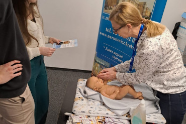 Widok z bok - Kobieta i mężczyzna stoją obok siebie, na przeciwko nich stoi Pani położna. Między nimi stoi stół, na którym znajduje się lalka - niemowlę.