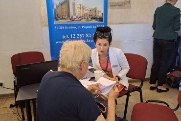 Pracownica szpitala rozmawia ze starszą kobietą. Obydwie osoby siedzą przy czarnym stoliku.