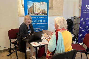 Pracownica szpitala rozmawia ze starszą kobietą. Obydwie osoby siedzą przy czarnym stoliku.