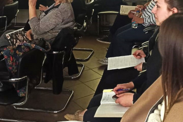 Słuchacze siedzą na krzesłach, bokiem do fotografa. Jedna z osób pije wodę.