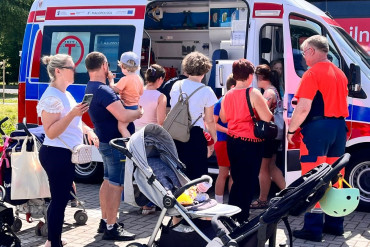 Zdjęcie przedstawia karetkę do której ustawiła się kolejka osób dorosłych z dziećmi