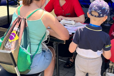 Swie kobiety siedzą przy stoliku, rozmawiając. Obok kobiety siedzącej tyłem do kamery stoi mały chłopiec.
