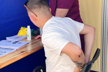 Kobieta - fizjoterapeutka stoi obok siedzącego mężczyzny, który trzyma ręce na dole pleców.