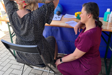 Na zdjęciu dwie kobiety. Fizjoterapeutka kuca obok siedzącej kobiety. Siedząca kobieta trzyma ręce z tyłu głowy ze splecionymi palcami.