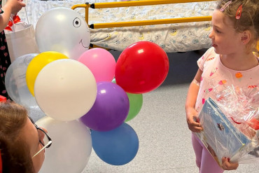 Kobieta kuca przed małą dziewczynką, trzymając w ręce balony. Dziewczynka trzyma zapakowany prezent.