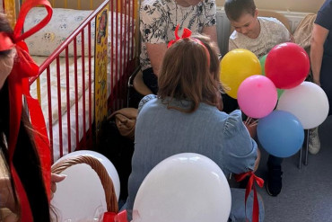 Chłopiec siedzi na krześle, obok siedzi mężczyzna. W ich kierunku kuca kobieta trzymająca kolorowe balony.
